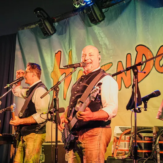 Zwei Männer treten auf der Bühne auf; einer singt in ein Mikrofon, während er Gitarre spielt, und der andere hält eine Gitarre. Scheinwerfer sind über ihnen, und ein Banner mit orangefarbenem Text lautet 'Hendon'.