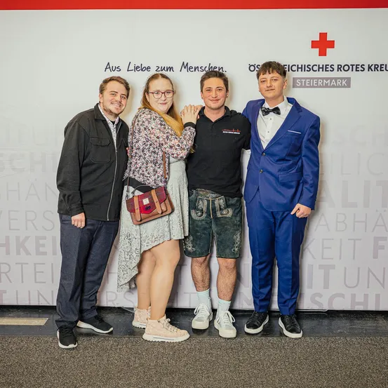 Vier Personen posieren für ein Foto vor einer Wand mit dem Text 'Aus Liebe zum Menschen.' Zwei Männer und eine Frau lächeln, während der vierte einen blauen Anzug trägt.