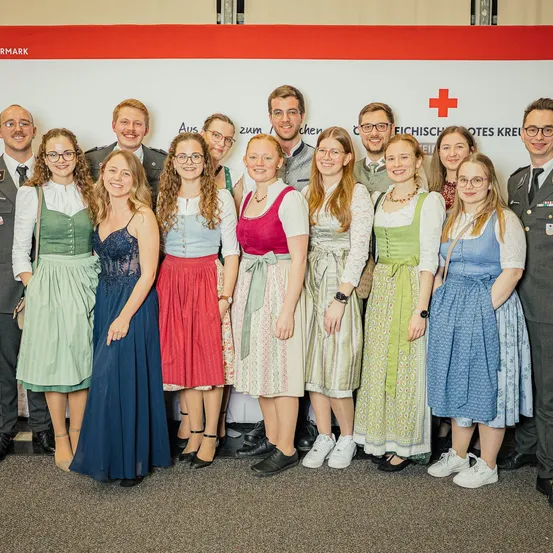 Eine Gruppe von Menschen in traditioneller deutscher Kleidung posiert für ein Foto vor einer Wand mit einem roten Kreuzschild und einem Banner mit weißem Text.