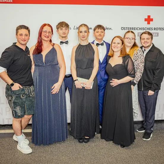 Eine Gruppe von sieben Personen posiert für ein Foto vor einer Wand mit dem Logo des Österreichischen Roten Kreuzes. Sie sind feierlich gekleidet, wobei zwei Frauen Abendkleider tragen.