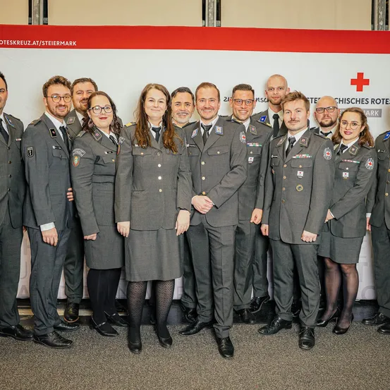 Eine Gruppe uniformierter Personen, wahrscheinlich aus einem militärischen oder Rettungsdienst, posiert für ein Foto vor einem weißen Hintergrund mit einem roten Kreuz.
