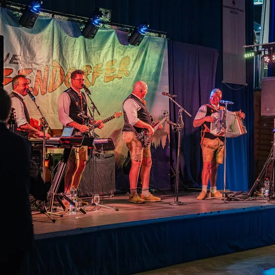 Eine Band aus vier Männern spielt auf der Bühne mit Musikinstrumenten unter einem Banner, das 'BENDORFER' steht. Ein Mann spielt Akkordeon, ein anderer Gitarre, während die anderen beiden Keyboard spielen.
