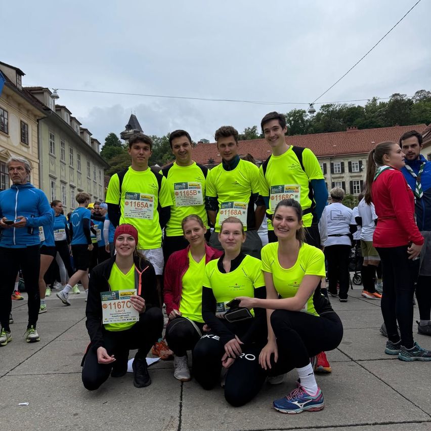 Eine Gruppe von Läufern in leuchtend gelben Shirts posiert für ein Foto mit Nummern auf der Brust vor einem Gebäude mit Bäumen und einem bewölkten Himmel.