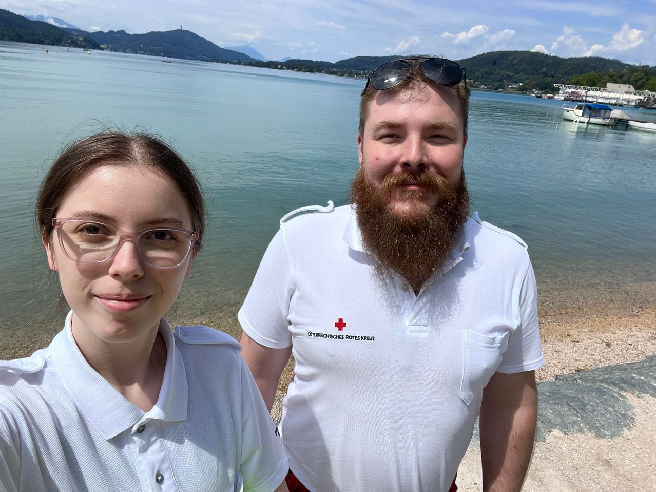 Zwei Menschen stehen am Strand in der Nähe eines ruhigen Meeres und lächeln. Der Mann hat einen Bart und eine Sonnenbrille auf dem Kopf, während die Frau eine Brille trägt. Sie tragen beide weiße Polo-Shirts mit dem Text 'Deutsches Rotes Kreuz'. In der Ferne gibt es Berge und ein Boot.