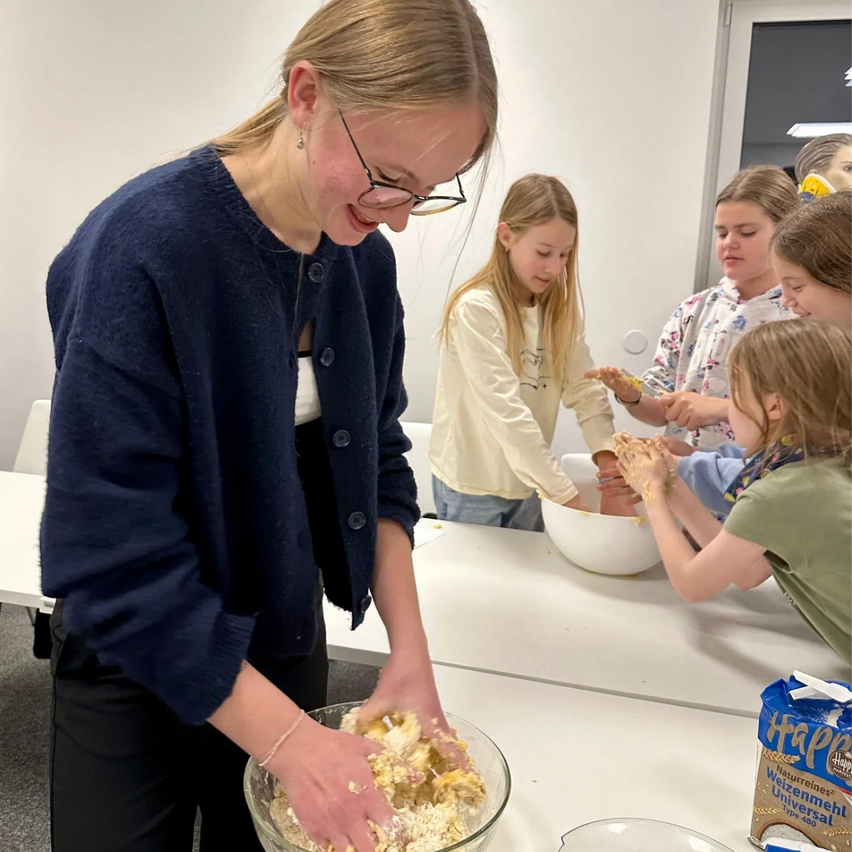 Eine Frau mit Brille und einem blauen Pullover führt eine Gruppe von Kindern in einer Küche an und zeigt ihnen, wie man Zutaten in einer Schüssel mischt. Die Kinder beobachten aufmerksam und folgen ihren Anweisungen.