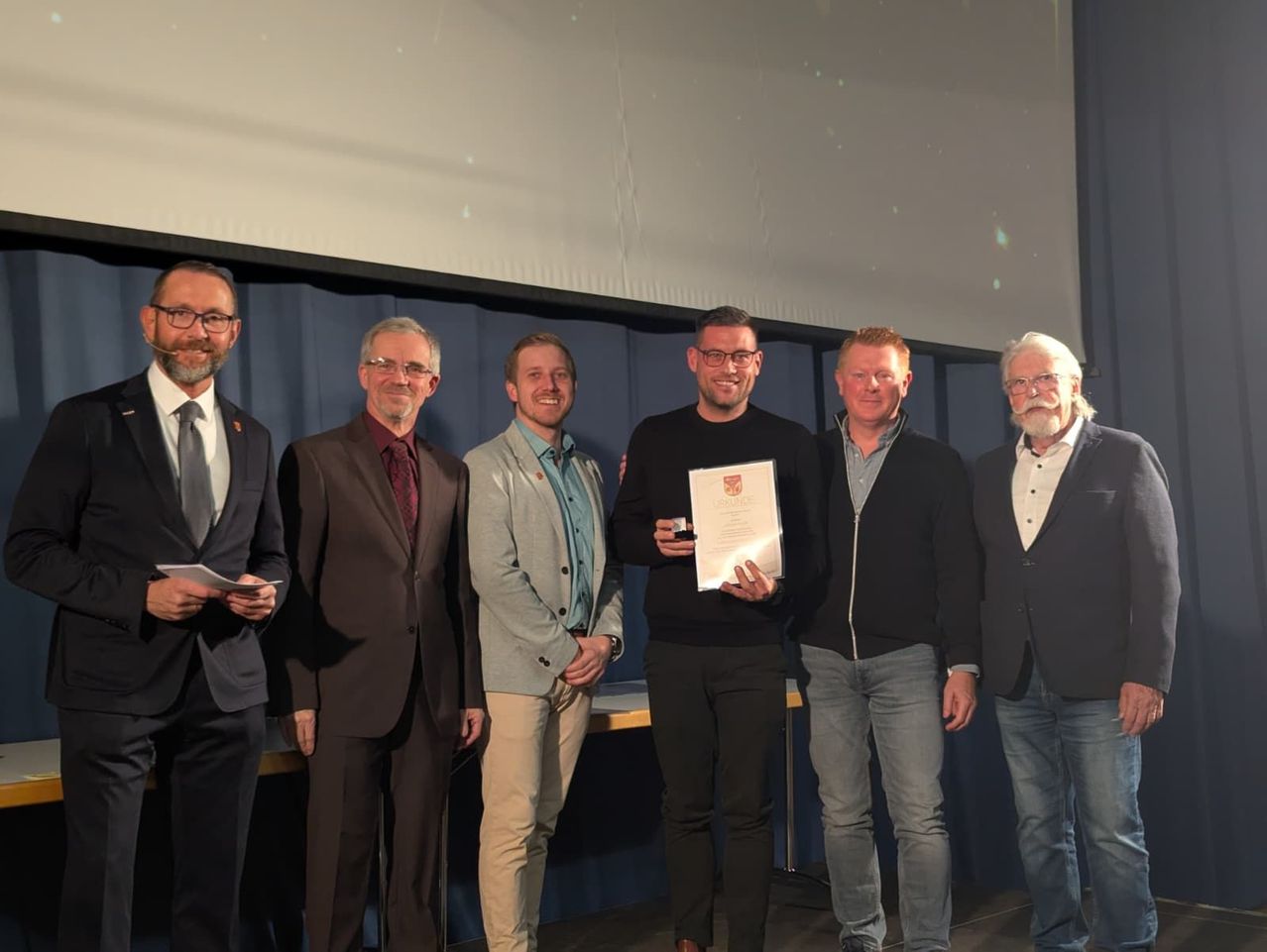 Six men in formal attire stand together, smiling. One man holds a certificate and a trophy. They are in front of a white screen.