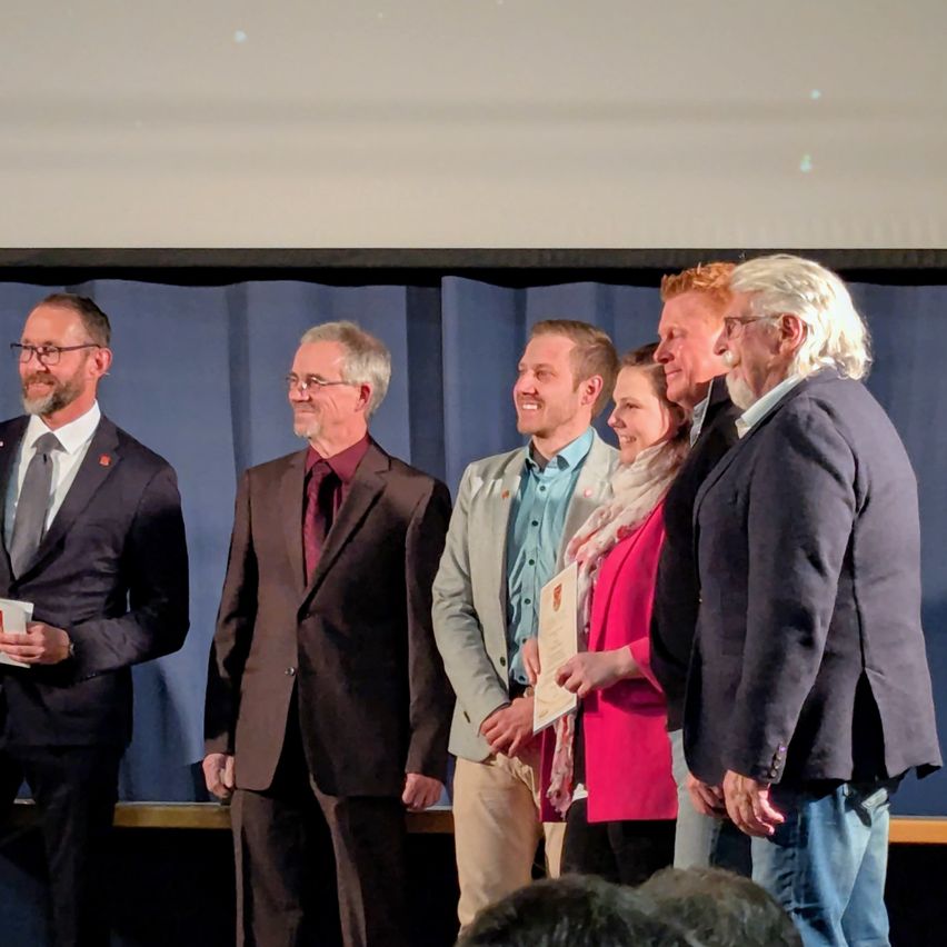 A group of people, including men and women, stand on a stage. They appear to be celebrating an achievement. One man holds a paper and a cup.