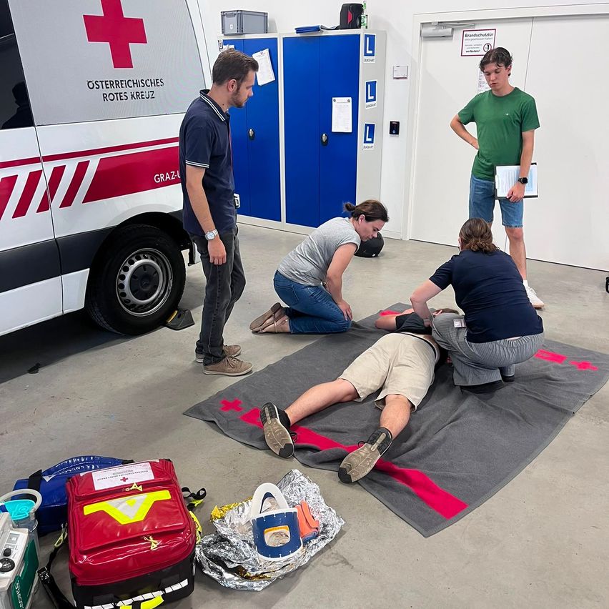A group of people in a garage, one person lying on a blanket, two people kneeling beside them, and two people standing. There is a red cross on the ambulance and a red cross on the blanket.