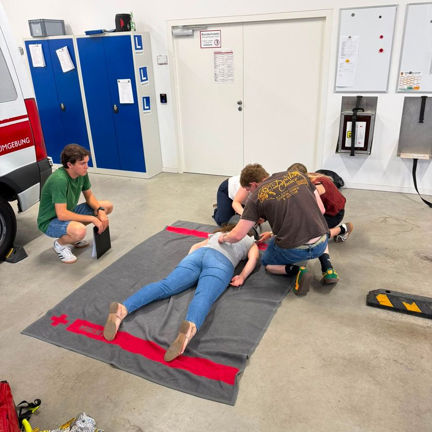 A group of individuals are practicing first aid on a woman lying on a mat in a garage. One person is kneeling and appears to be performing CPR while others are observing.