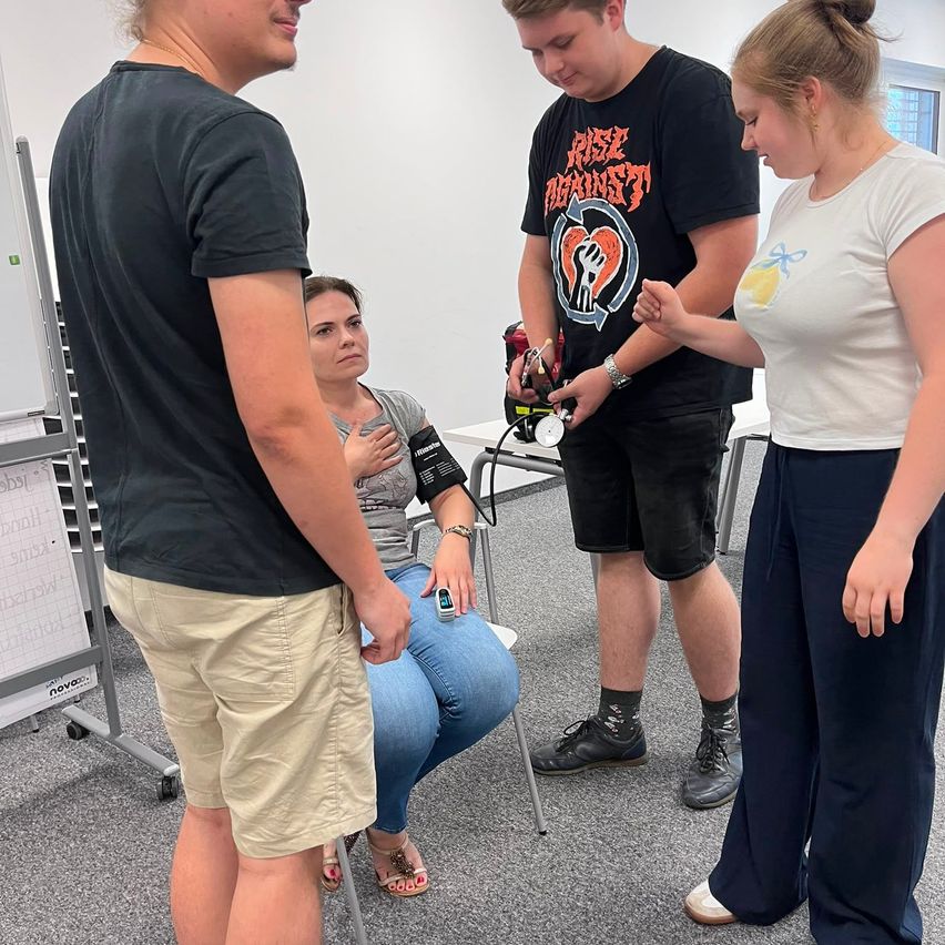 Three individuals are gathered in a room, with one person seated on a chair while another person stands beside her, holding a blood pressure monitor. The third person stands in front of them, looking at the monitor.