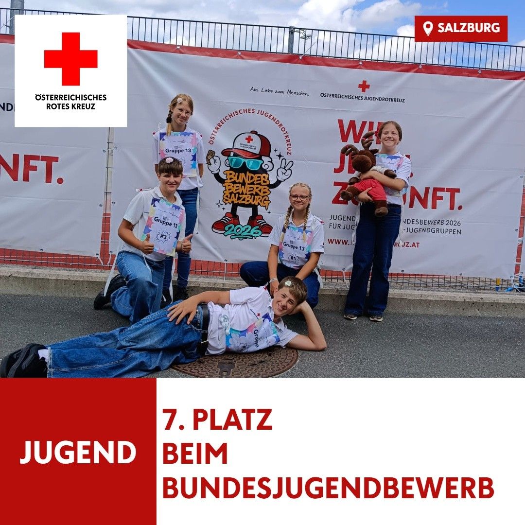 Fünf junge Personen, die weiße T-Shirts mit roten Kreuzen tragen, posieren für ein Foto vor einem Banner mit dem Text 'Jugend beim Bundesjugendbewerb'. Sie halten Urkunden in den Händen und einer von ihnen hält ein Kuscheltier.
