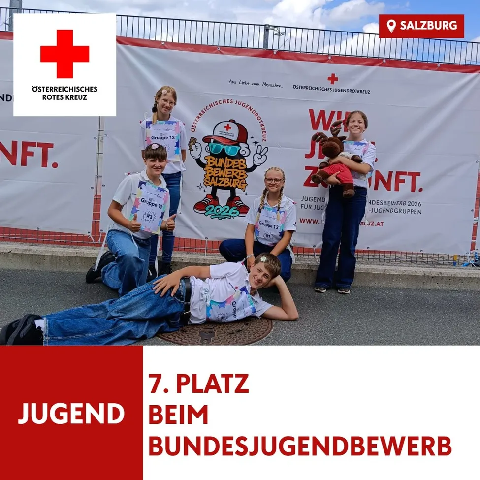 Fünf junge Personen, die weiße T-Shirts mit roten Kreuzen tragen, posieren für ein Foto vor einem Banner mit dem Text 'Jugend beim Bundesjugendbewerb'. Sie halten Urkunden in den Händen und einer von ihnen hält ein Kuscheltier.