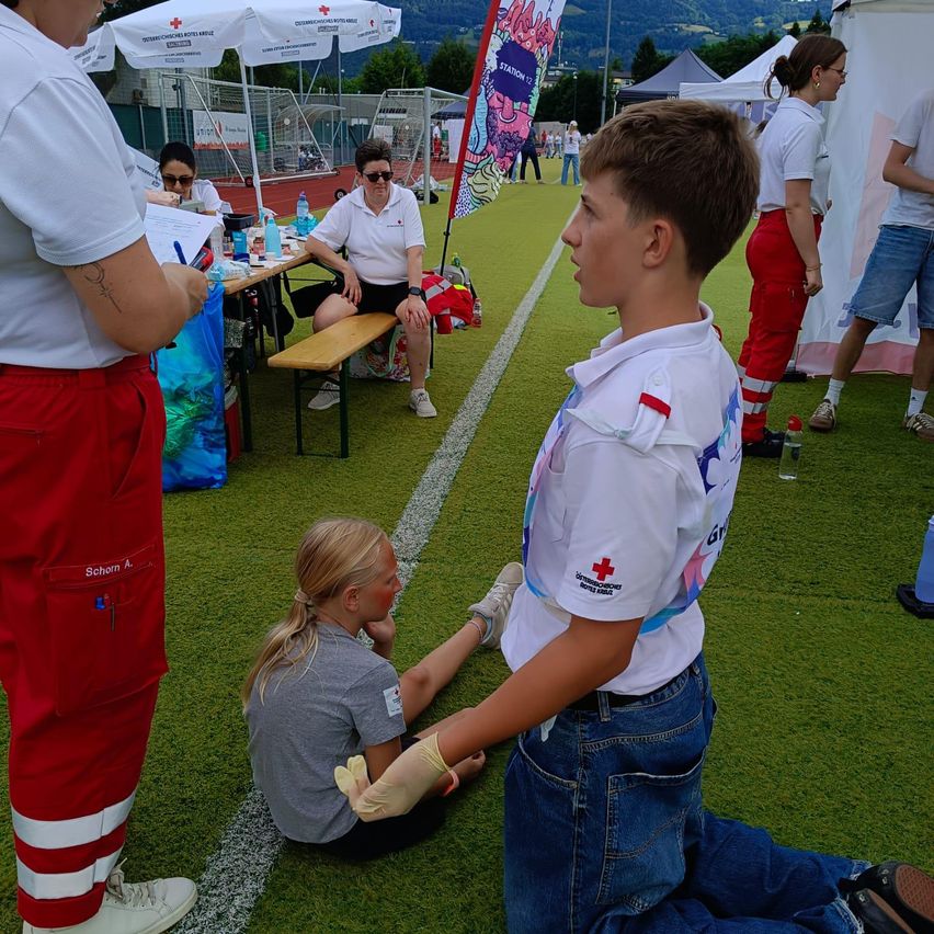Ein Junge in einem weißen Hemd mit rotem Kreuz kniet auf einem Feld, möglicherweise bei einem Erste-Hilfe-Training. Eine Mädchen sitzt in der Nähe mit verbundener Hand.