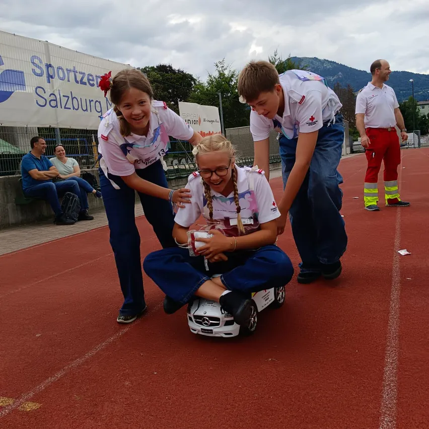 Drei Kinder in weißen Uniformen sind auf einer Rennstrecke, eines sitzt auf einem Spielzeugauto, ein anderes hilft ihr, und ein Mann in roten Hosen steht in der Nähe.