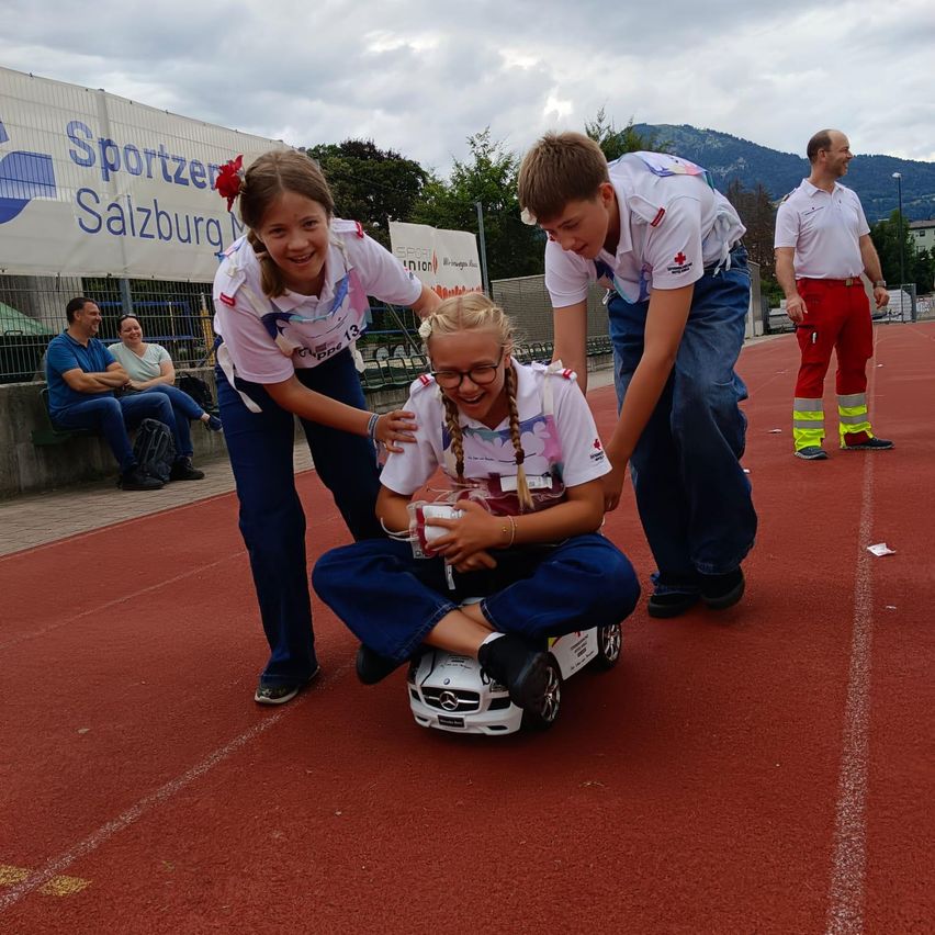 Drei Kinder in weißen Uniformen sind auf einer Rennstrecke, eines sitzt auf einem Spielzeugauto, ein anderes hilft ihr, und ein Mann in roten Hosen steht in der Nähe.