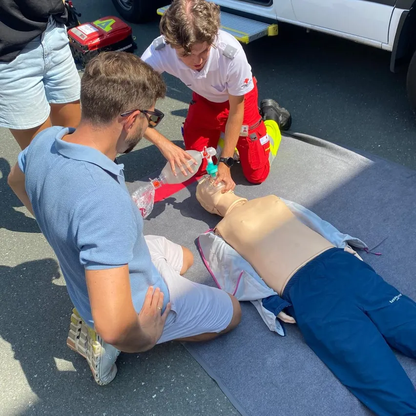 Zwei Männer üben CPR an einem Dummiesarg auf dem Boden. Ein Mann kniet, der andere sitzt und hält eine Flasche. Eine dritte Person steht hinter ihnen.