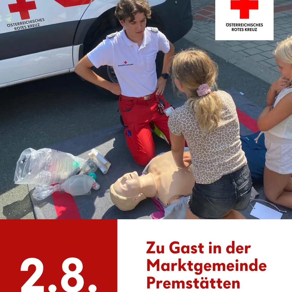 Ein junger Mann in roter und weißer Uniform kniet neben einer Puppe und demonstriert möglicherweise CPR. Ein Mädchen beobachtet ihn, und ein weiteres Mädchen sitzt in der Nähe. Die Szene spielt sich im Freien ab, mit einem roten Kreuz-Ambulanz im Hintergrund.