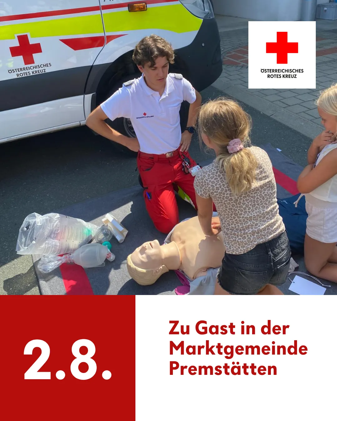 Ein junger Mann in roter und weißer Uniform kniet neben einer Puppe und demonstriert möglicherweise CPR. Ein Mädchen beobachtet ihn, und ein weiteres Mädchen sitzt in der Nähe. Die Szene spielt sich im Freien ab, mit einem roten Kreuz-Ambulanz im Hintergrund.