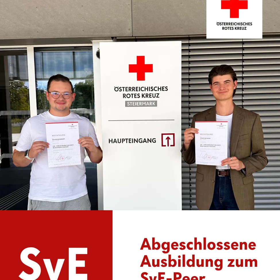 Zwei Männer stehen vor einem Gebäude und halten Urkunden in den Händen. Der Mann auf der linken Seite trägt eine Brille und ein weißes T-Shirt, während der Mann auf der rechten Seite eine braune Jacke und einen Ring trägt. Hinter ihnen ist ein Banner mit dem Text 'Haupteingang' und einem roten Kreuz-Logo angebracht.