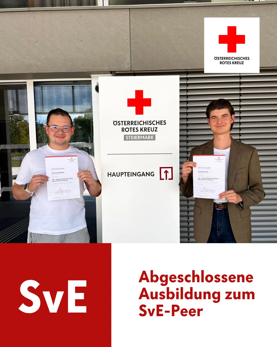 Zwei Männer stehen vor einem Gebäude und halten Urkunden in den Händen. Der Mann auf der linken Seite trägt eine Brille und ein weißes T-Shirt, während der Mann auf der rechten Seite eine braune Jacke und einen Ring trägt. Hinter ihnen ist ein Banner mit dem Text 'Haupteingang' und einem roten Kreuz-Logo angebracht.