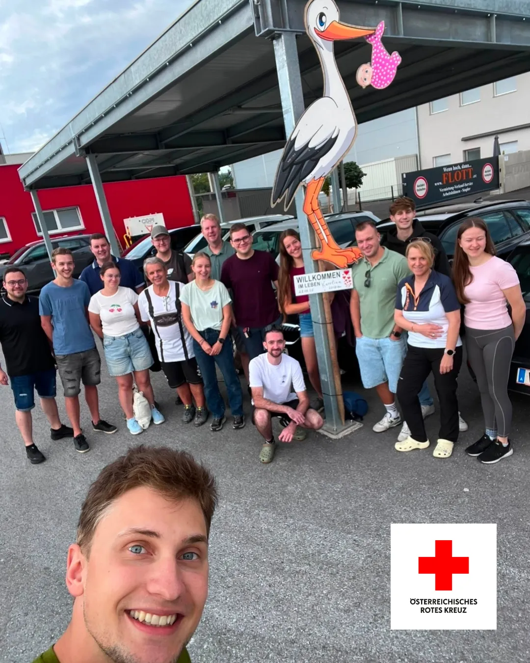 Eine Gruppe von Menschen posiert für ein Foto auf einem Parkplatz mit einer Storchstatue und einem Schild, das 'Willkommen' sagt.
