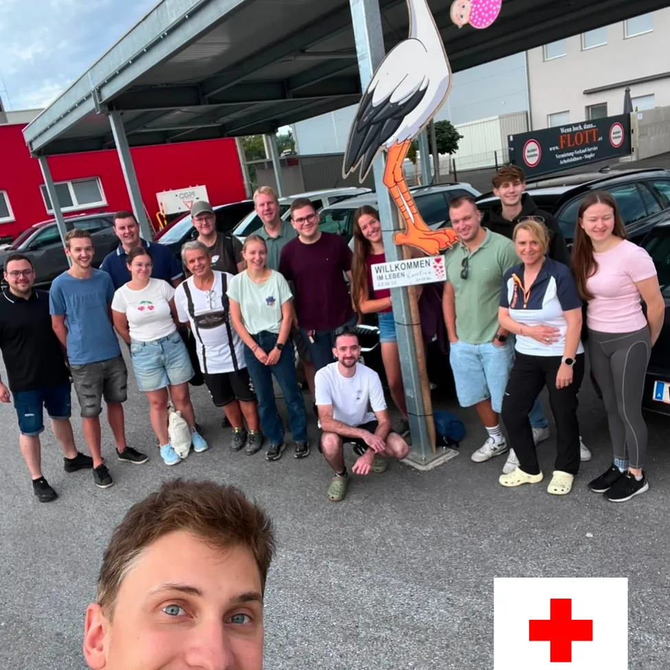 Eine Gruppe von Menschen posiert für ein Foto auf einem Parkplatz mit einer Storchstatue und einem Schild, das 'Willkommen' sagt.