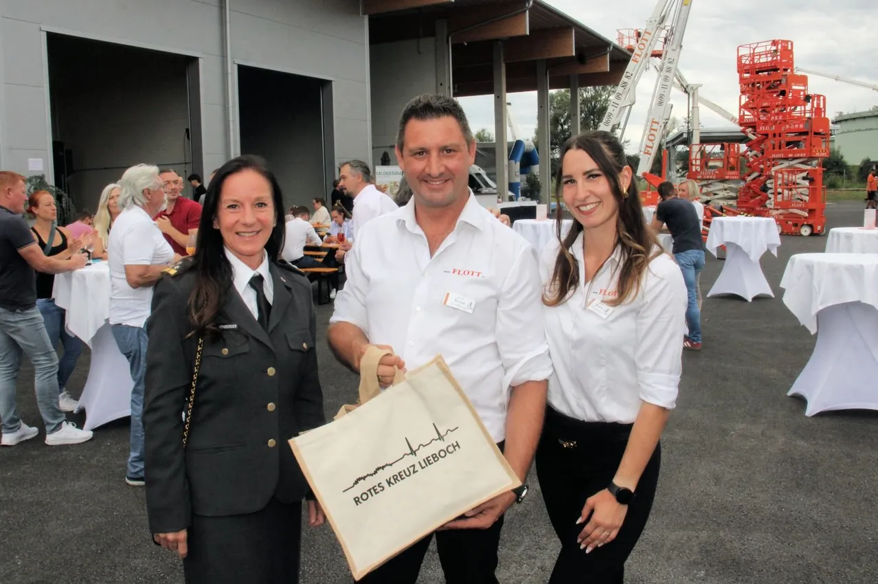 Drei Personen stehen lächelnd draußen. Ein Mann hält einen Beutel mit der Aufschrift 'Rotes Kreuz Lieboch'. Eine Frau in Uniform steht links von ihm, und eine andere Frau in einem weißen Hemd steht rechts von ihm. Hinter ihnen sitzen Leute an Tischen, und ein Gebäude mit einer offenen Tür ist im Hintergrund.
