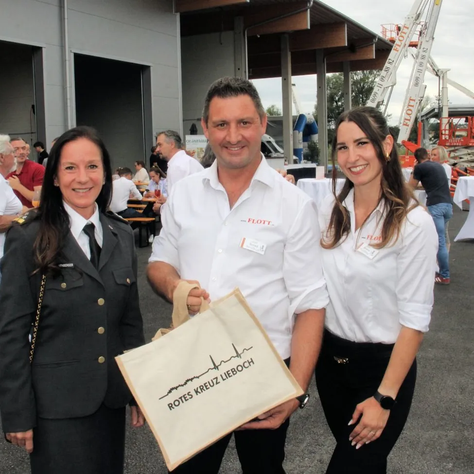 Drei Personen stehen lächelnd draußen. Ein Mann hält einen Beutel mit der Aufschrift 'Rotes Kreuz Lieboch'. Eine Frau in Uniform steht links von ihm, und eine andere Frau in einem weißen Hemd steht rechts von ihm. Hinter ihnen sitzen Leute an Tischen, und ein Gebäude mit einer offenen Tür ist im Hintergrund.
