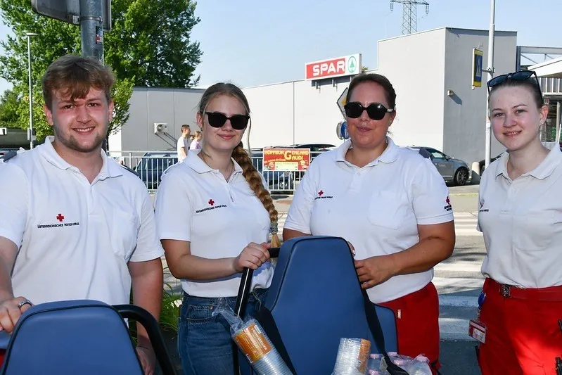 Vier Personen in weißen Poloshirts mit rotem Kreuz stehen draußen. Die Frau in der Mitte hält eine blaue Tasche. Dahinter ist ein Gebäude mit einem Spar-Schild zu sehen.