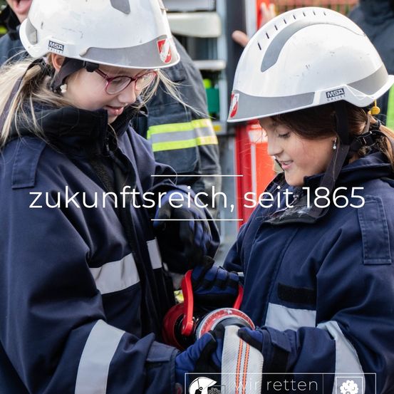 Zwei Frauen in Helmen schauen sich an und halten ein rotes Seil. Sie tragen Uniformen mit reflektierenden Streifen. Das Bild hat den Text Zukunftsreich, seit 1865.
