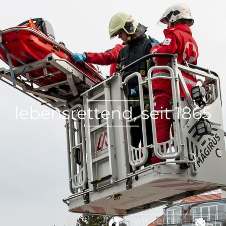 Zwei Feuerwehrleute stehen auf einer Leiter mit einer roten Trage, die auf einer erhöhten Plattform steht, und tragen rote Uniformen und Helme. Die Plattform hat ein Logo und den Text 'Magirus'.