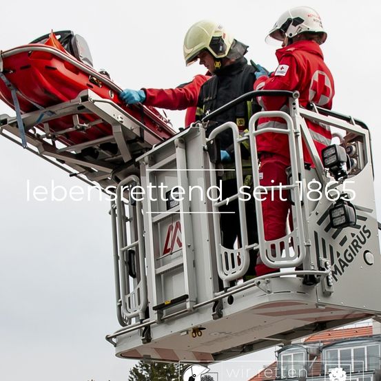 Zwei Feuerwehrleute stehen auf einer Leiter mit einer roten Trage, die auf einer erhöhten Plattform steht, und tragen rote Uniformen und Helme. Die Plattform hat ein Logo und den Text 'Magirus'.