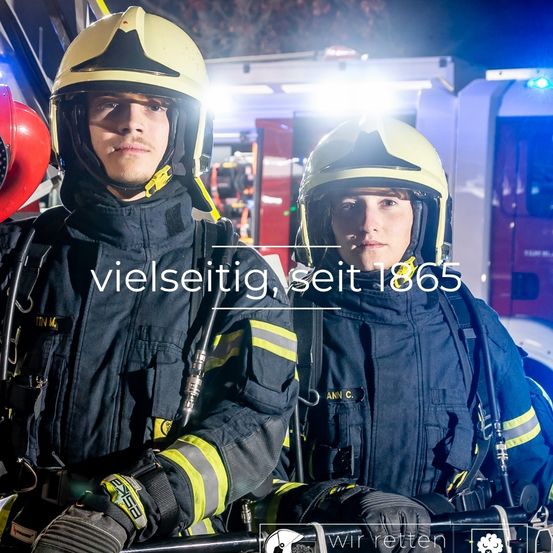 Zwei Feuerwehrleute in Uniform stehen vor einem Feuerwehrwagen. Sie tragen beide Helme und Handschuhe. Der Text lautet Vielseitig, seit 1865.