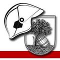 Ein heraldisches Emblem zeigt einen Feuerwehrhelm, einen Baum und ein Fass. Der Baum wächst aus dem Fass, das mit Schutt gefüllt ist. Der Schild ist mit einem roten Streifen umrandet.