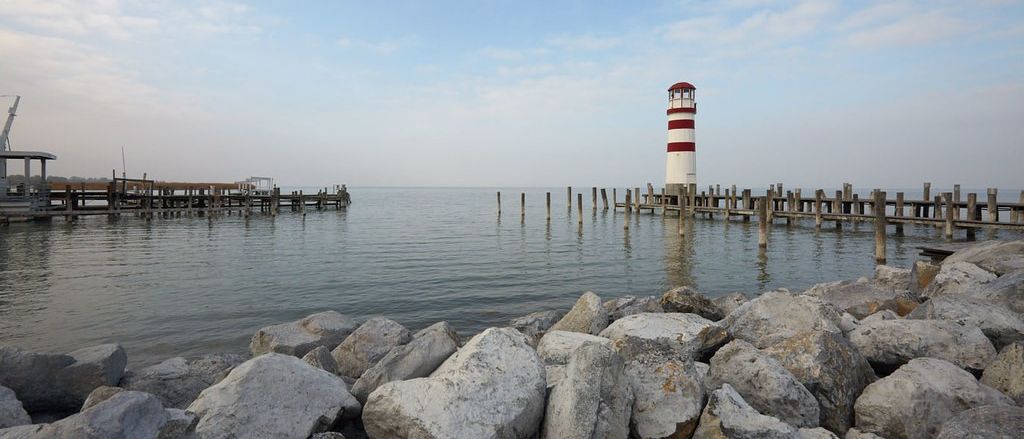 Ein Leuchtturm steht auf einem Steg, der sich in ein ruhiges Meer mit felsigen Wellenbrechern im Vordergrund erstreckt. Der Himmel ist blau mit verstreuten Wolken.
