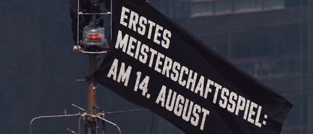 Zwei maskierte Personen stehen auf einem Pfosten und halten ein Banner mit der Aufschrift 'Erstes Meisterschaftsspiel: Am 14. August' vor dem Hintergrund eines hohen Gebäudes.