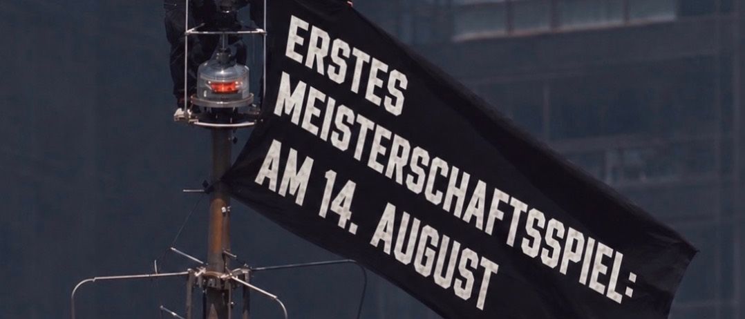 Zwei maskierte Personen stehen auf einem Pfosten und halten ein Banner mit der Aufschrift 'Erstes Meisterschaftsspiel: Am 14. August' vor dem Hintergrund eines hohen Gebäudes.