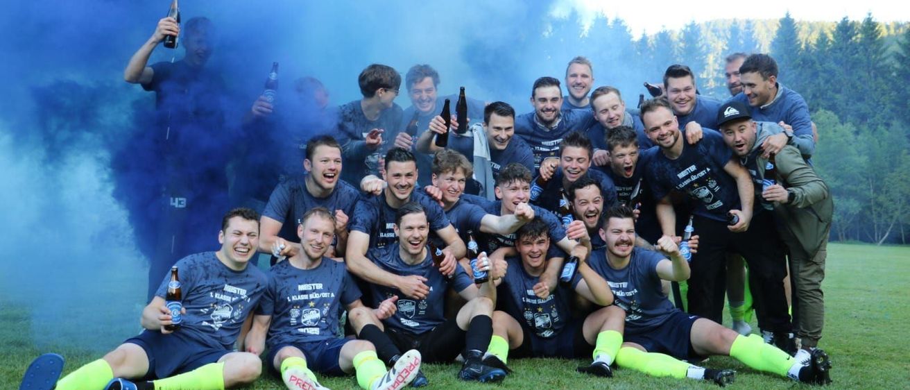 Ein Fußballteam feiert einen Sieg auf einem Feld, umgeben von blauem Rauch und hält Bierflaschen. Sie tragen passende blaue Trikots mit weißen Buchstaben und Mützen.
