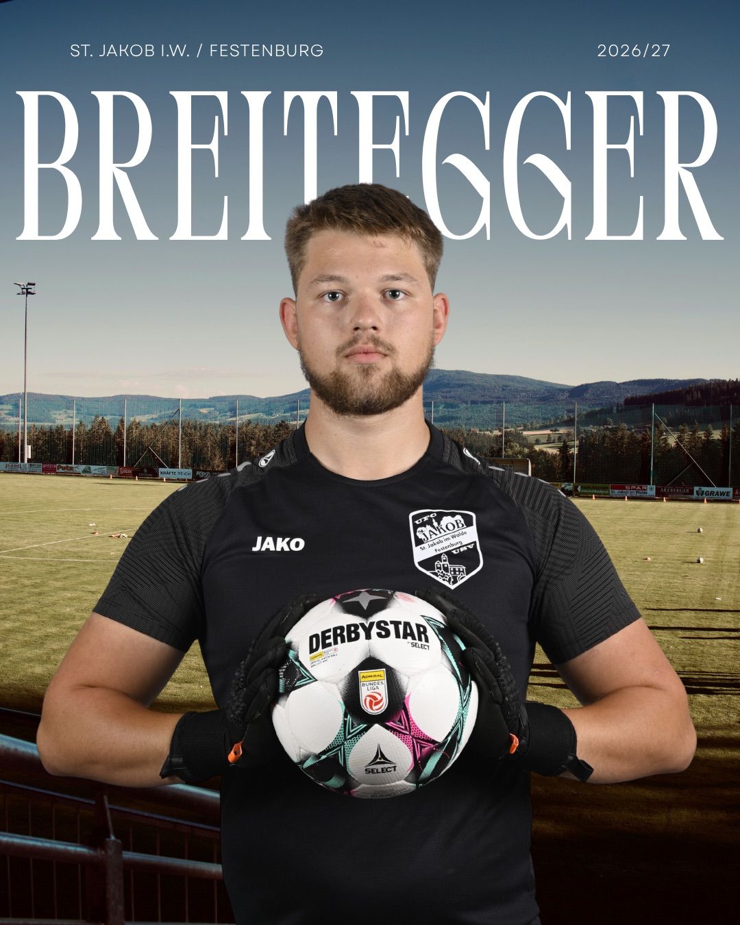 A man in a black soccer jersey with the word Jako on it is holding a soccer ball. The background shows a soccer field with mountains in the distance.