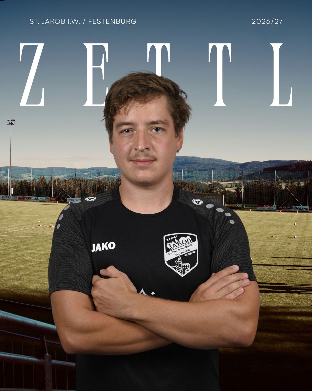 A young man stands in a soccer field wearing a black jersey with the word Jako and a logo on the chest. He is smiling and posing with his arms crossed. Behind him is a blurry background of a soccer field with a fence and mountains.