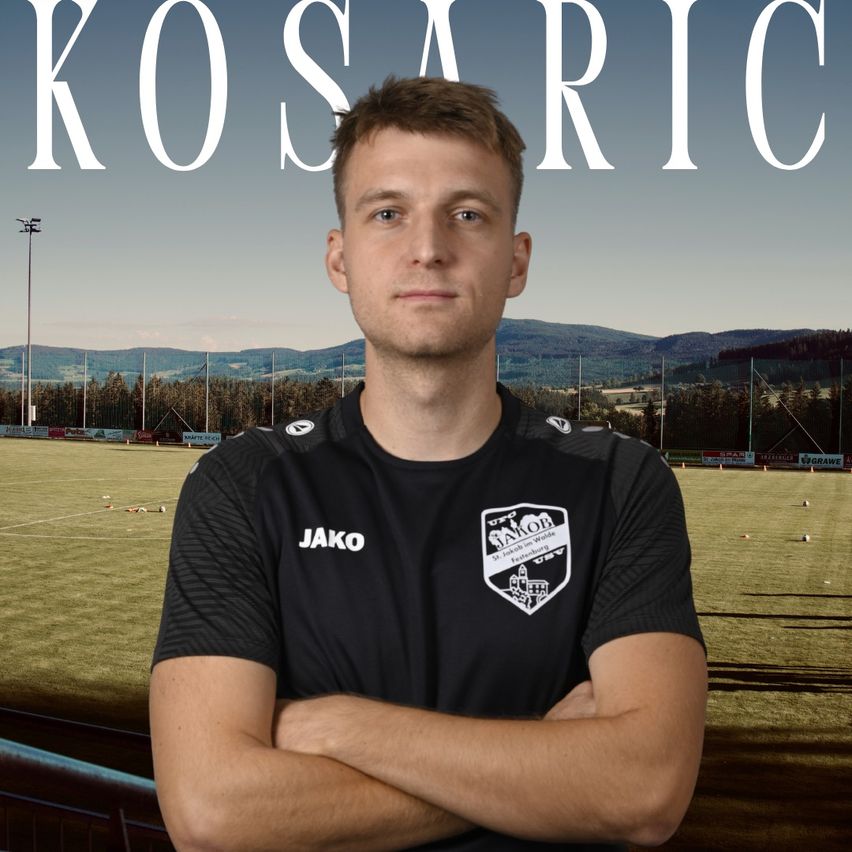A man stands in front of a soccer field with the word Kosaric in the background. He wears a black jersey with the word Jako on it.