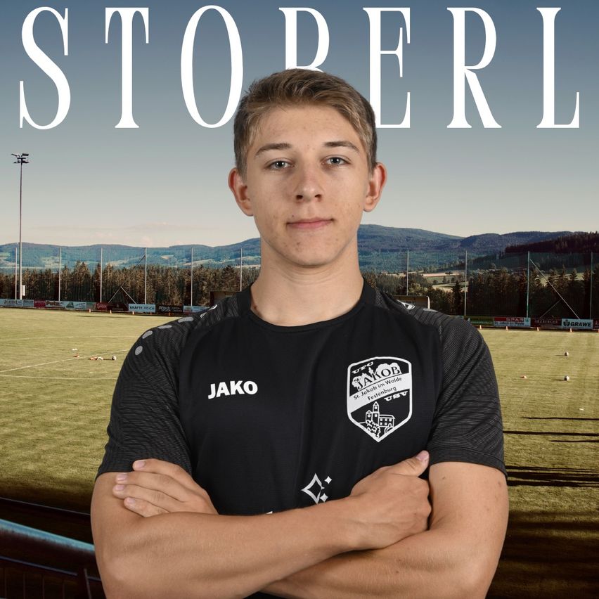 A young man stands on a soccer field with his arms crossed, wearing a black jersey with the word 'Jako' and a logo. The background features a grassy field with a fence and mountains. The word 'STOPERL' is displayed prominently above him.
