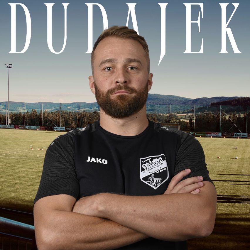 A man with a beard stands on a soccer field, wearing a black shirt with the word 'Jako' and a logo. He has his arms crossed and is looking to the side. Behind him is a blurry background with a field, trees, and mountains. The word 'Dudajek' is written in large white letters above him.