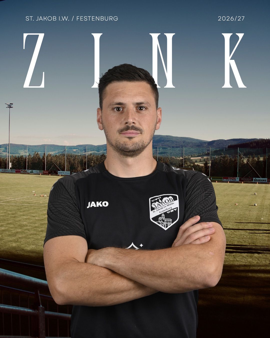 A man in a black soccer jersey with the word 'Jako' on it stands with his arms crossed in front of a soccer field.