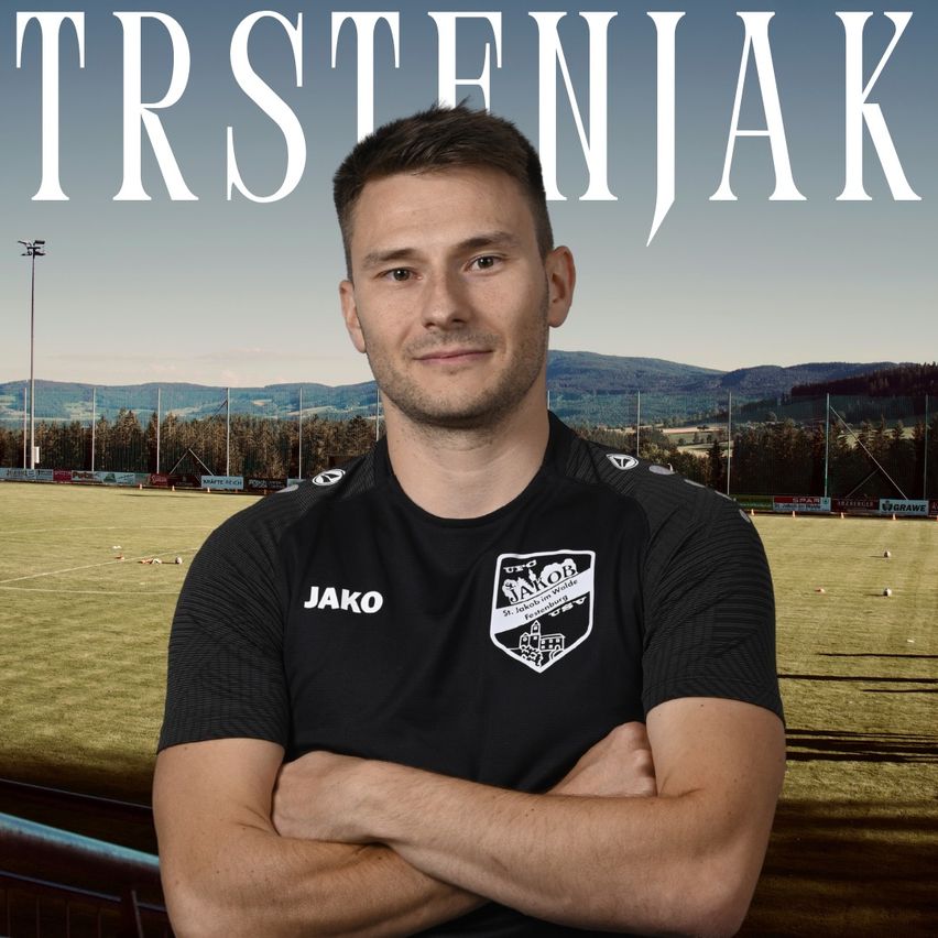 A man in a black soccer jersey with the word 'Jako' on it is standing on a soccer field with his arms crossed. The background shows a scenic view of mountains and a clear sky.