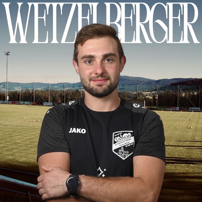 A man stands on a soccer field with a smiling face, wearing a black Jako shirt with a logo on the left chest. Behind him is a blurry background of a soccer field with a mountain range.