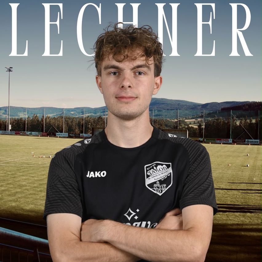 A man with curly hair stands in front of a soccer field, wearing a black jersey with the word Jako on it. The background shows a soccer field with a mountain range in the distance.