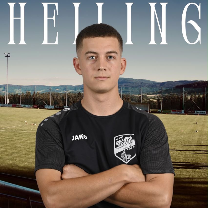 A young man in a black soccer jersey with the word Jako on it stands with his arms crossed in front of a soccer field.