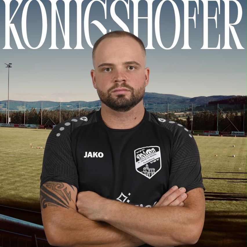 A man in a black sports jersey with the word Jako on it, standing in front of a soccer field.