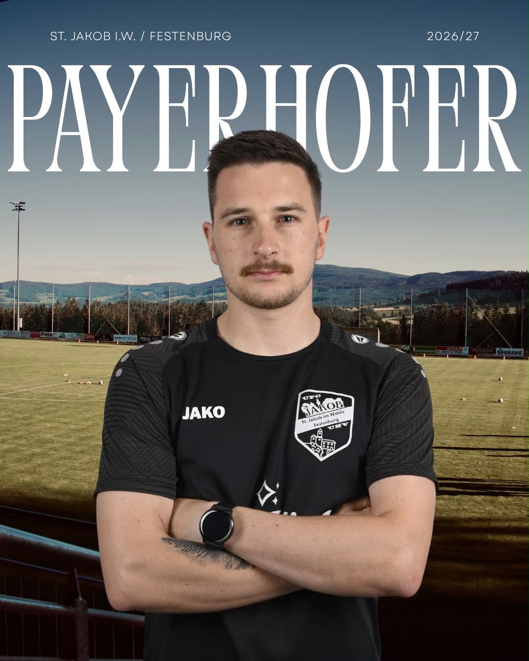 A man with a mustache and beard stands in front of a soccer field, wearing a black soccer jersey with the word 'Jako' on it. He has his arms crossed and is looking directly at the camera. The background shows a soccer field with a blurry view of trees and mountains.